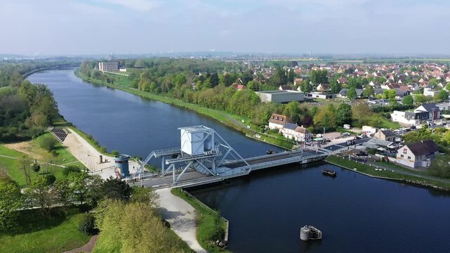 Aerial view of Pegasus Bridge spanning a calm river, surrounded by lush greenery and a residential neighborhood in Normandy, France. Bright, clear spring day with scenic landscape and historic