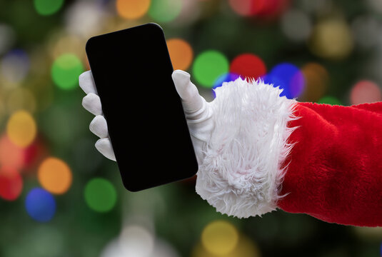 Cropped shot of an Santa Claus hand showing a black mobile phone screen at home with christmas tree