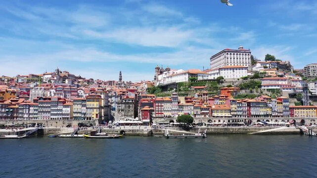 Vibrant traditional buildings line the riverfront in the historic center of Porto, Portugal, under a clear blue sky. The cityscape features colorful facades and terraced architecture overlooking the