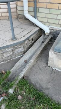 Rainwater downpipe draining into concrete gutter, apartment building base shows brick wall, concrete channel lined with grass.