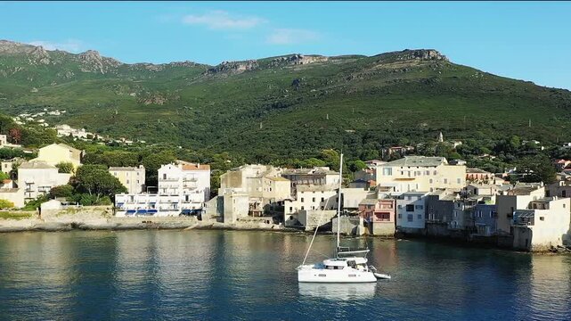 A white sailboat floats on calm water near the picturesque waterfront houses of Erbalunga village, with lush green hills in the background, Corsica, France.
