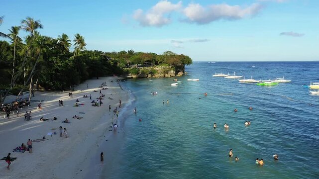 Aerial view of a tropical beach with people relaxing on the sand and swimming in clear blue water, with traditional boats anchored offshore in Panglao, Philippines.