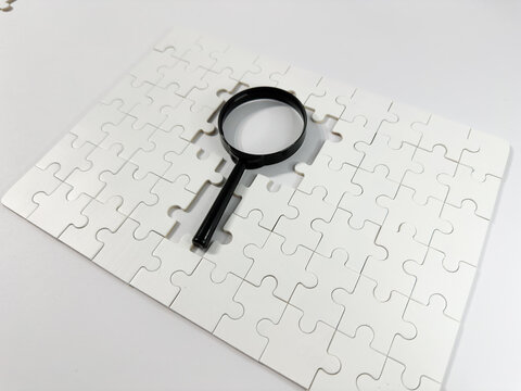 A magnifying glass rests on a blank white jigsaw puzzle, symbolizing investigation, problem-solving, analysis, seeking answers, and the process of uncovering missing information or solutions.