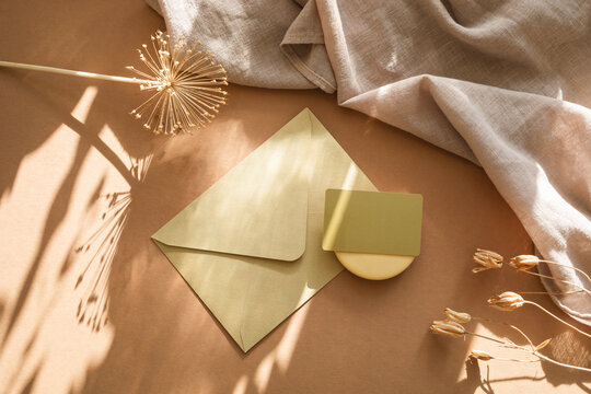 Overhead view of a blank business card on an envelope, a rustic vase with dried flowers, candle and napkin on a  table in sunlight
