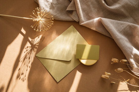 Overhead view of a blank business card on an envelope, a rustic vase with dried flowers, candle and napkin on a  table in sunlight