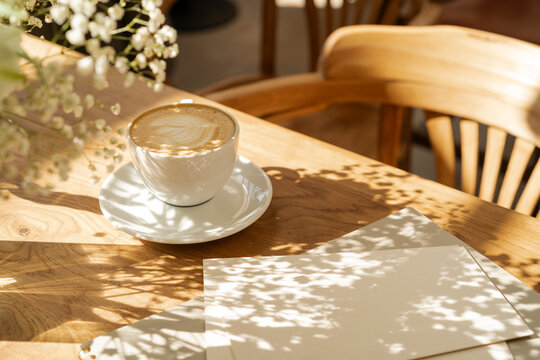 Close-up of a cup of coffee, blank pieces of paper and floral shadows  on a wooden table on a sunny morning