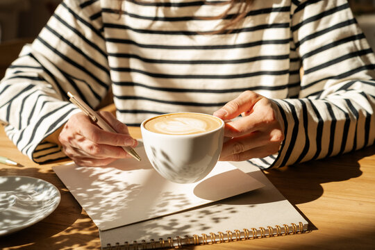 Close-up of a Woman in a striped top sitting at wooden table about to write on a blank piece of paper while drinking a latte coffee on a sunny morning
