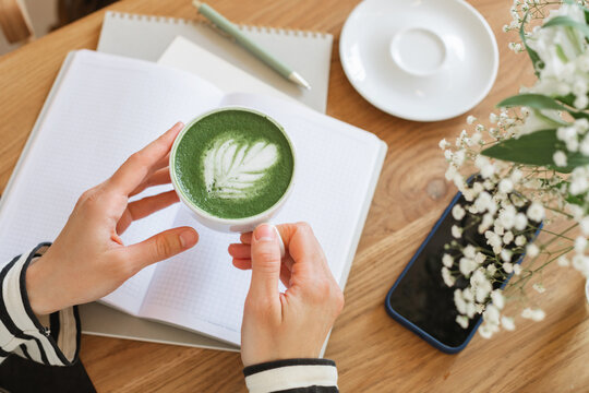 Overhead view of a woman sitting at a table with an open notebook and mobile phone drinking a hot matcha latte