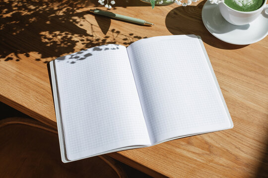 Close-up overhead view of an open notebook with blank pages and a hot matcha latte drink on a wooden table with floral shadows