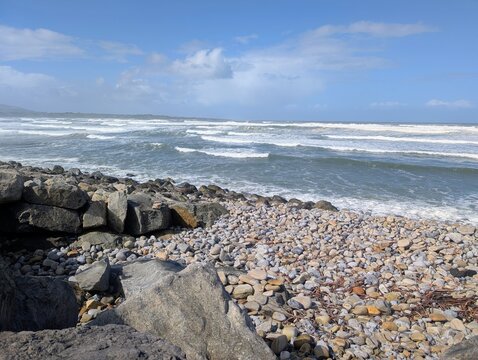 Powerful Atlantic waves roll onto the rocky shoreline at Strandhill Beach in County Sligo, Ireland. A dramatic coastal scene featuring textured pebbles and rugged boulders. 
