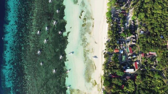 Drone shot of clear turquoise waters, white sandy beach, and traditional boats along the coast of Panglao Island, Philippines. Lush greenery and small buildings line the shoreline.