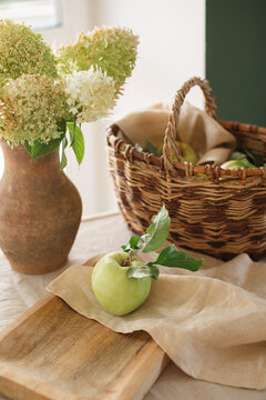 Bunch of fresh hydrangeas in a rustic clay jug on a table with a basket and an apple on a wooden chopping board with a napkin