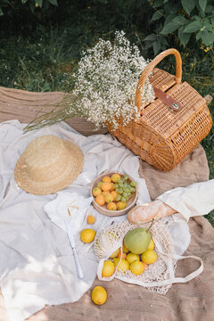 Overhead view of a wicker basket, bunch of baby's breath flowers, straw hat, fresh baguette and assorted fruit on a picnic blanket in a summer garden