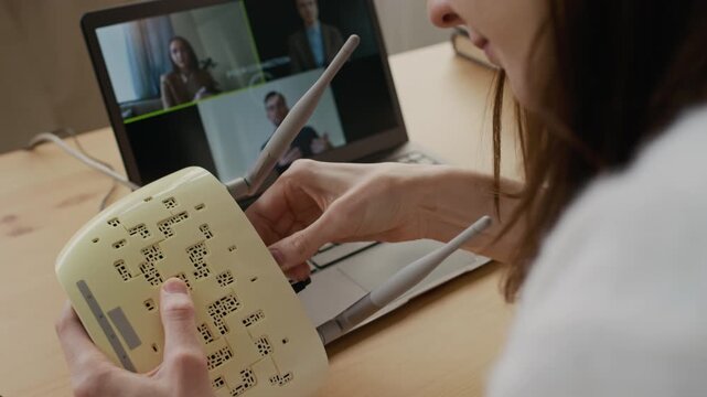 Over-shoulder shot of woman trying to reset internet connection after losing signal during business video call, plugging cables and pressing buttons on router, looking at frozen image on laptop screen
