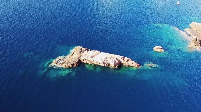 Aerial view of rocky islets surrounded by clear blue water in the Calanques de Piana, Corsica. The sunlight reveals underwater rock formations and vibrant turquoise hues.