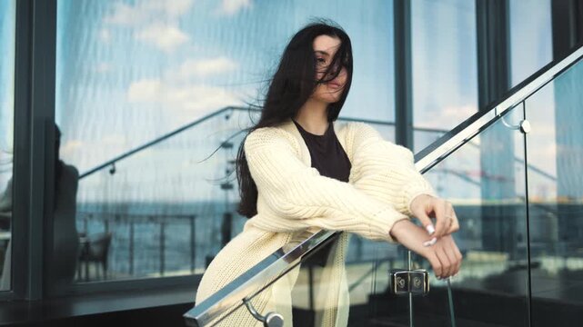A stylish young woman leans on a glass railing of a modern terrace, looking at the city view. Urban, lifestyle, fashion, elegance, city, morning, comfort, serenity, architecture, business, office