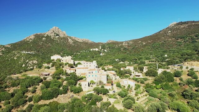 Scenic aerial view of the traditional village of Pigna nestled among lush green hills and rugged mountains in Corsica, France, under a clear blue sky.