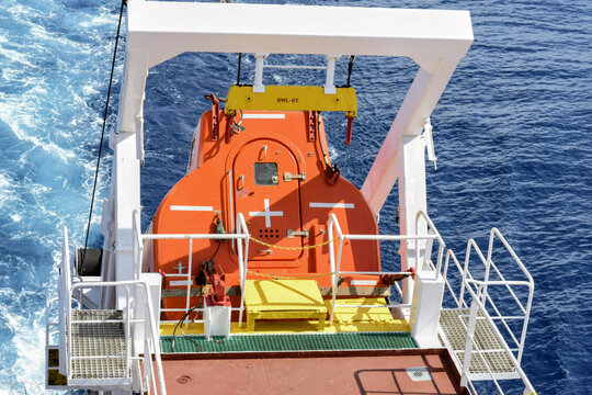 an enclosed lifeboat hangs from a ship davit above open water, with ocean wake visible below showing motion and readiness for emergency deployment
