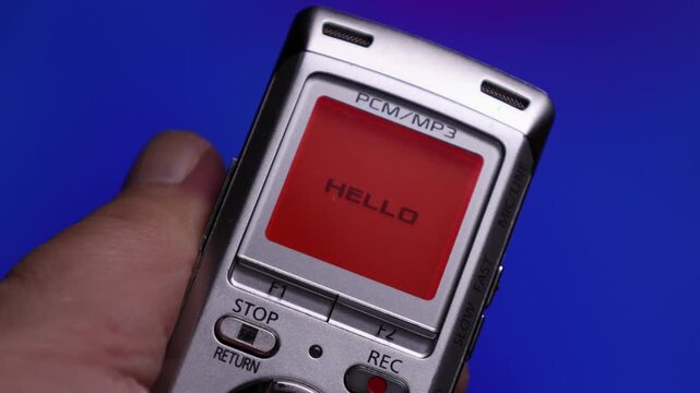 Portable digital voice recorder held in a persons hand against a blue background. The device features a small screen with labels indicating PCMMP3 recording formats, control buttons including Stop,.