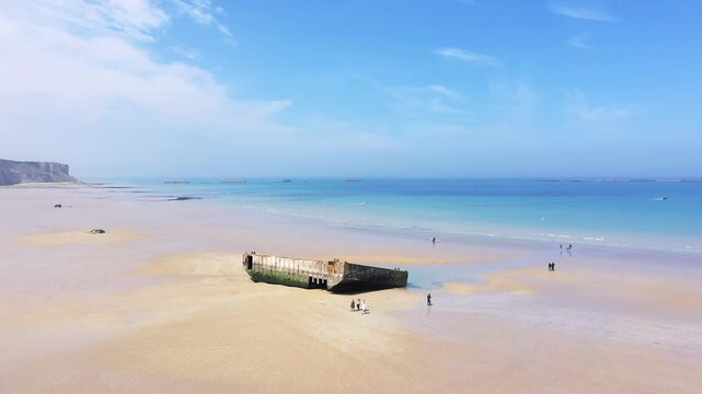 Wide sandy beach at Arromanches les Bains, Normandy, featuring remnants of World War II Mulberry harbors under a clear blue sky. Tranquil seascape with scattered visitors and calm waters.