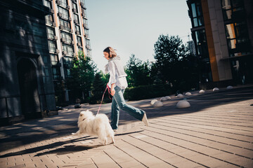 Young woman walking a fluffy white dog on a sunny urban plaza between modern apartment buildings, enjoying a leisurely city stroll © deagreez