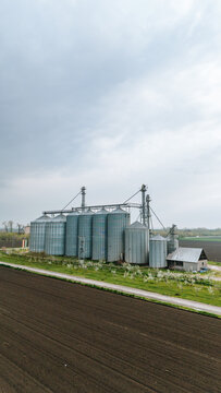 Aerial view of the towering silver grain elevators standing guard over the dark, tilled earth under a vast, muted sky, Sremska Mitrovica, Vojvodina, Serbia.
