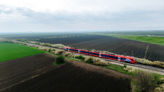 Aerial view of a vibrant red and blue train cuts through the landscape, contrasting against the dark ploughed fields and verdant green patches, Sremska Mitrovica, Vojvodina, Serbia.