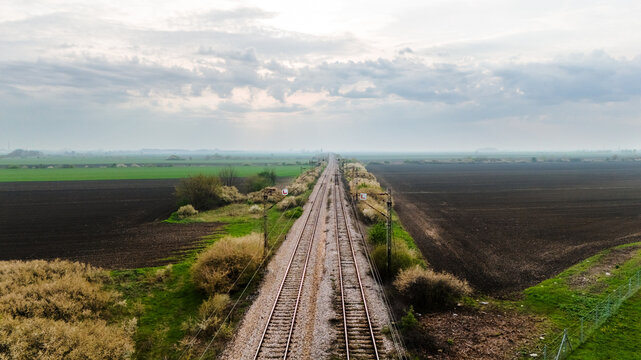 Aerial view of railway tracks cutting through contrasting fields of vibrant green and dark soil under a soft, diffused sky, Sremska Mitrovica, Vojvodina, Serbia.