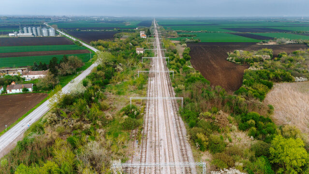Aerial view of a long railway track cutting through green fields and trees, with a cluster of silos visible in the distance, Sremska Mitrovica, Vojvodina, Serbia.
