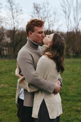 Fototapeta premium Romantic couple embraces and kisses outdoors in knit sweaters, candid lifestyle shot in a park, warm natural light, affectionate moment, serene field setting, intimate connection.