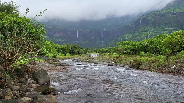 A tiny spring trickles gently in the foreground while a powerful waterfall thunders behind, set against lush green monsoon mountains, creating a serene yet dramatic natural spectacle.