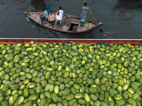 Dhaka, Bangladesh - 04 April 2026: Aerial view of a boat laden with watermelons, its green and yellow hues contrasting with the dark water, as passengers observe the vibrant harvest.