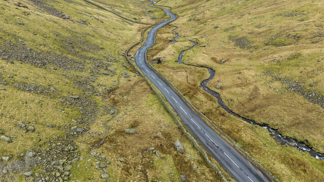 Aerial view of a winding road cutting through the rugged, grassy landscape, contrasting with the dark asphalt and the meandering stream, England, United Kingdom.