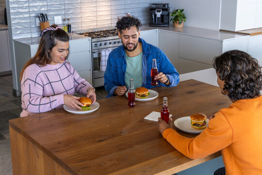 Three friends in casual wear sitting at kitchen island holding burgers and sipping drinks