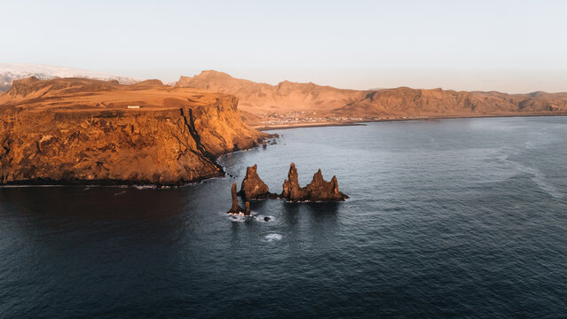 Aerial view of basalt sea stacks rising sharply from the dark ocean waters, framed by rugged cliffs under the soft glow of dawn, Reynisdrangar, Iceland.