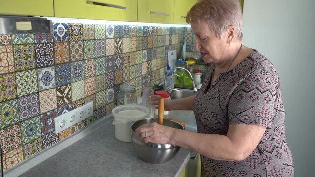 Grandmother pouring milk into a bowl. Elderly woman preparing dough at home in the kitchen. Kneading with a pestle. Cook food and talk