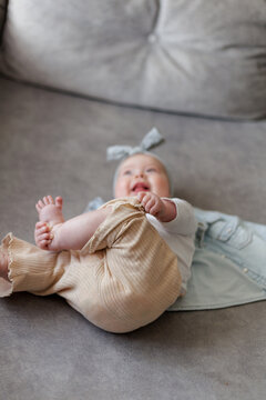 Joyful baby girl with bow headband playing with her feet while lying on a sofa