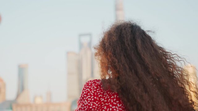 City skyline woman facing towers with flowing curly hair and glasses, morning glow and distant skyscrapers, panoramic vista, soft breeze moving hair, travel and discovery mood, roles include