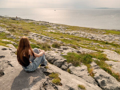 Woman sits on rocky cliff, gazing out at ocean. Her long hair flows freely in the coastal breeze. She appears calm, contemplative, deeply connected to nature. Burren, Ireland, Travel and tourism.
