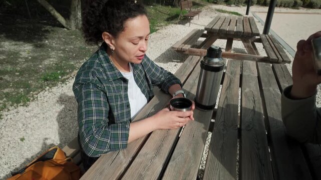Sharing a Warm Drink: Woman Pours Coffee from a Thermos at an Outdoor Picnic Table.