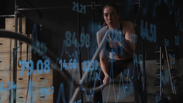 Woman gripping battle ropes in gym, making waves, blue data graphs crossing frame covering subject