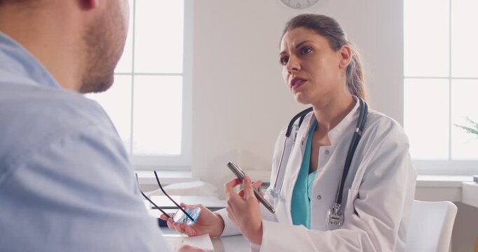 Serious doctor or nurse talks to a patient in a hospital, discussing medical exam results during a visit. Conversation highlights healthcare, examination, and professional medicare assessment.