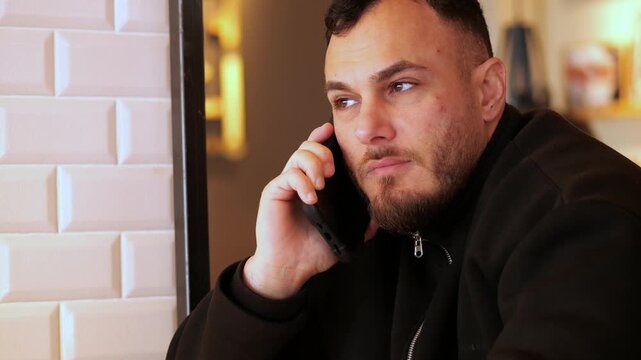 A medium shot of a young man with a beard, wearing a black jacket, engaged in
a phone call while sitting in a modern cafe. A glass mug of herbal tea with a
visible tea bag is on the wooden table 