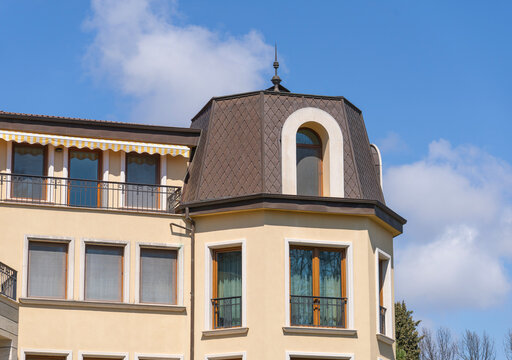 Elegant facade of a classic European-style building featuring a brown mansard roof, arched windows, and decorative balconies against a bright blue sky with soft white clouds