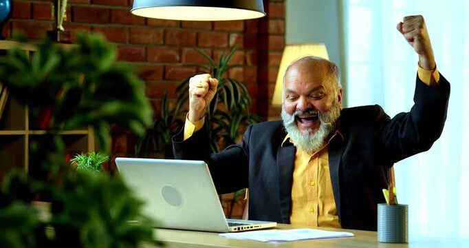 Indian senior businessman or corporate executive showing a successful gesture, such as a fist pump or confident smile, while working on his laptop at a desk in a cozy office with moody lighting.