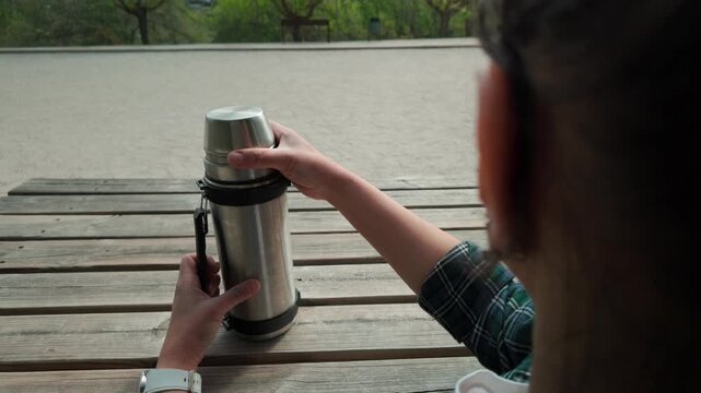 Woman Closes Thermos Bottle on a Wooden Bench Outdoors