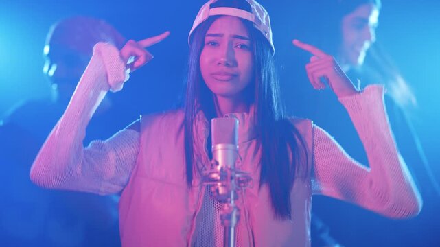 Talented female rapper singing and dancing in front of a microphone with her band in a studio with colored lights for a music video
