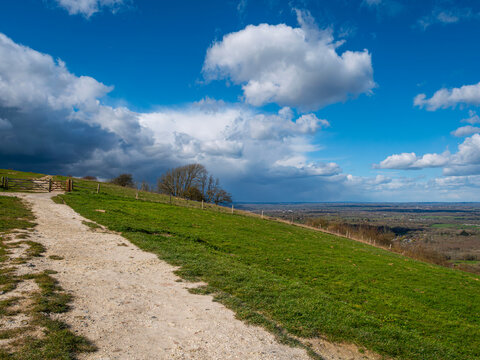 A public footpath leads to a wooden gate and fence along the South Downs Way at Ditchling Beacon, Sussex, on a green hillside overlooking a scenic countryside view. Blue sky, white and dark clouds.