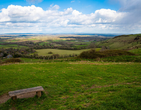 A wooden bench seat on a green hillside beside the South Downs Way public footpath at Ditchling Beacon, Sussex, overlooking a scenic countryside view of the Sussex Downs. Blue sky with clouds.