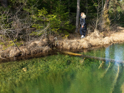 Beautiful blonde woman standing by the crystal clear emerald waters of a forest spring lake in Estonia during early spring